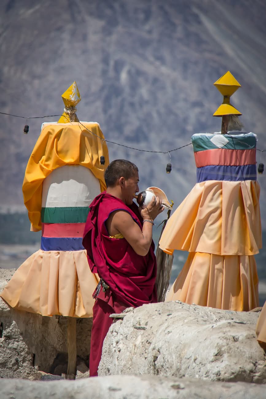 Moine bouddhiste soufflant dans une conque rituelle entre deux stupas colorés
